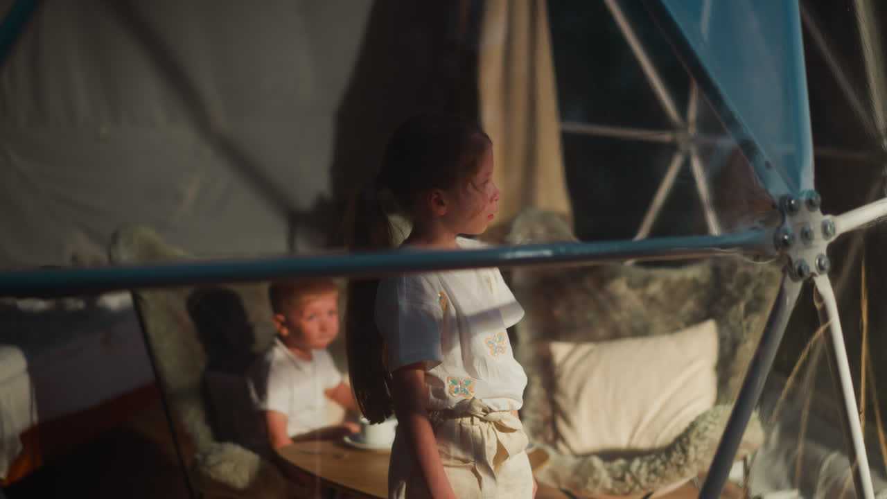 Little children in spherical glamping hut. Girl looks through panoramic window while brother rests in armchair. Kids spend time at night camping
