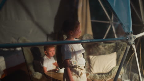 niños pequeños en una cabaña de glamping esférica. la niña mira a través de la ventana panorámica mientras el hermano descansa en el sillón. los niños pasan la noche acampando