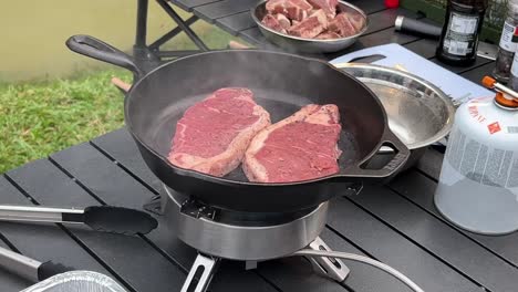 two cuts of steak sizzling on hot cast iron frying pan at outdoor camping site environment during daytime, close up shot