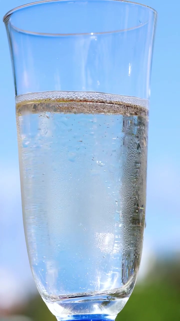 Un flûte da champagne trasparente riempito di spumante è tenuto all'aperto contro un cielo azzurro brillante, con bollicine visibili che salgono alla luce naturale del sole.
