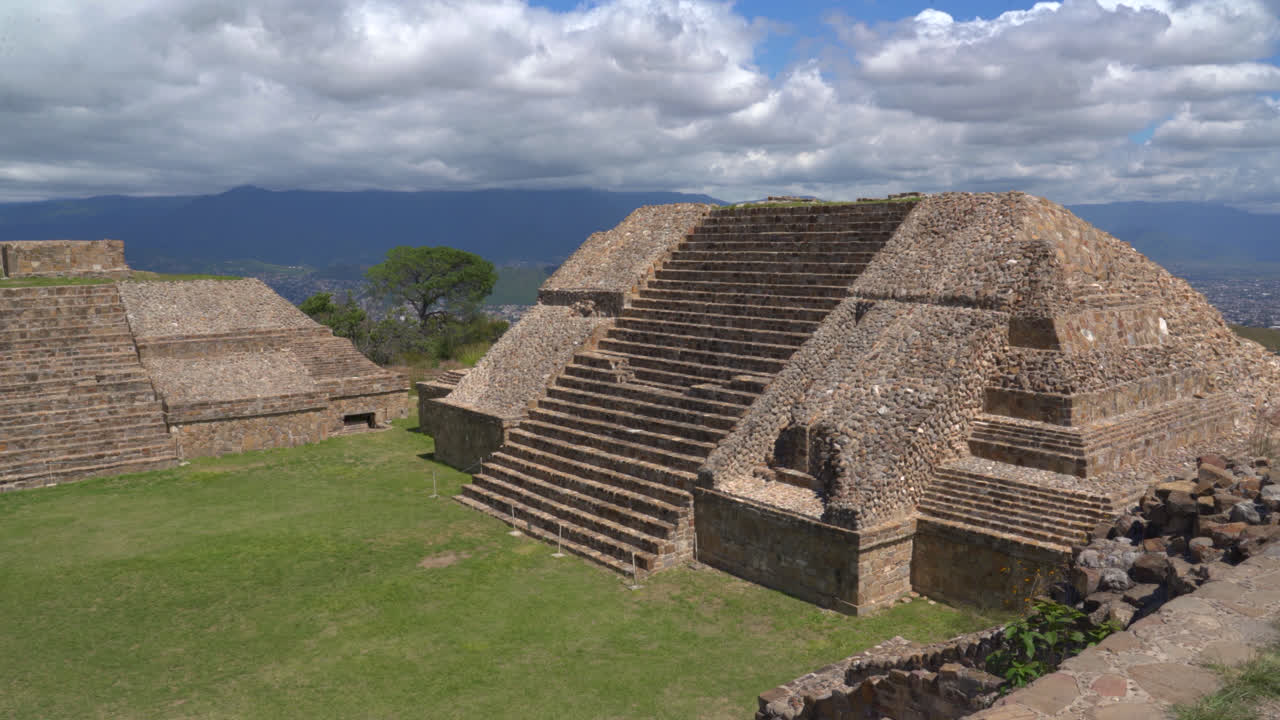 Monte Alban stairs and temple ruins stand across high ridge with stone silhouettes and warm valley light, establishing wide pan left