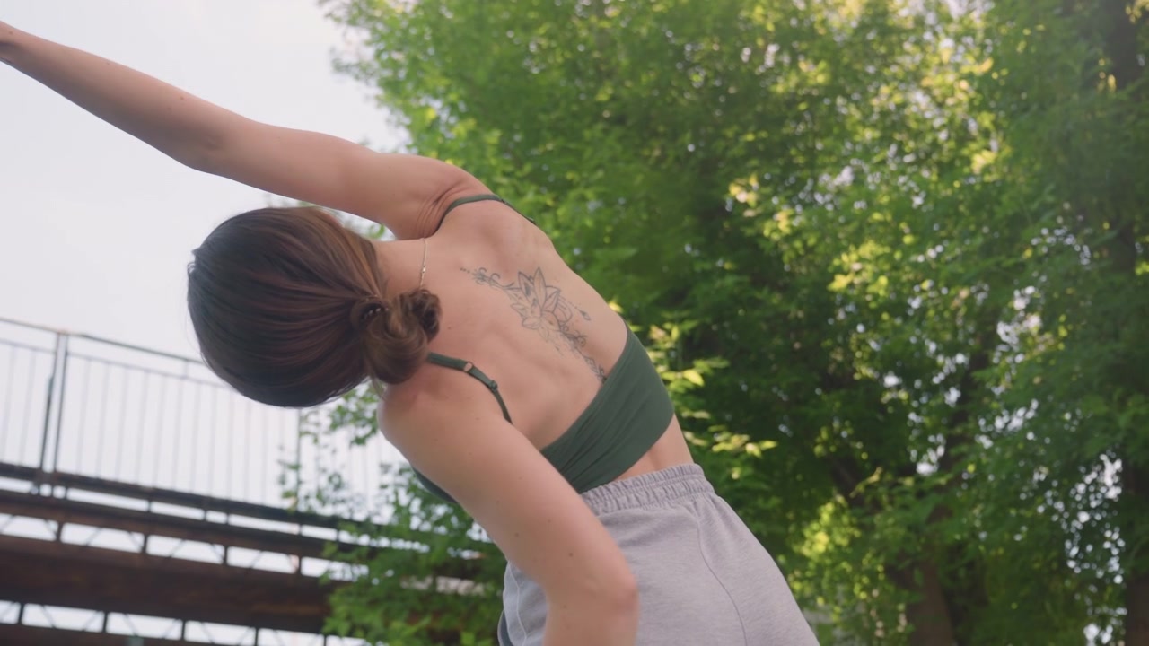 Close up of woman performing stretching exercises outdoors with her back to camera wearing athletic clothing and displaying visible tattoos. subject moves through various arm stretches