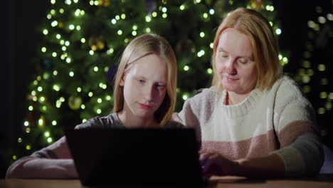 mamá e hija están mirando la pantalla de la computadora portátil juntos. sentados frente a las luces borrosas del árbol de navidad