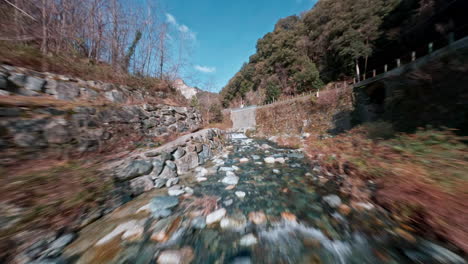 Vista-De-Drones-FPV-De-Un-Río-Pintoresco-Que-Fluye-A-Través-De-Una-Zona-Boscosa-Rocosa-Bajo-Un-Cielo-Azul-Brillante