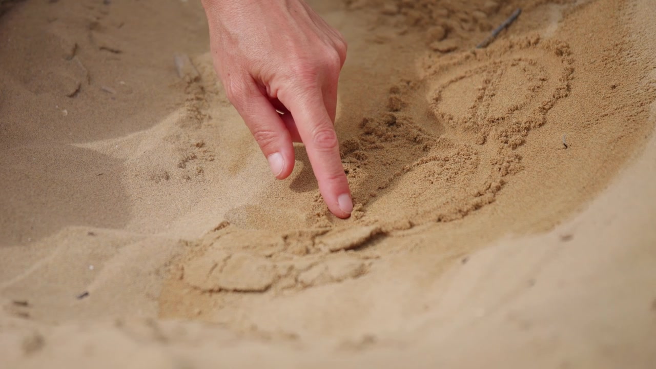 Närbild av en person som ritar former direkt i strandsanden med ett finger, skapar tillfälliga mönster under en avkopplande stund utomhus