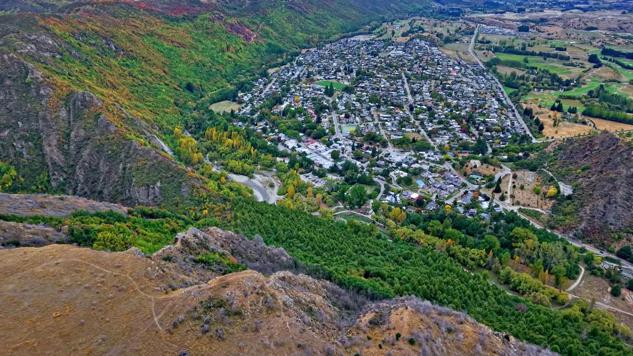 Aerial drone flies toward Arrowtown from German Hill, capturing scenic Central Otago hills, town buildings, and the surrounding lush landscape in New Zealand