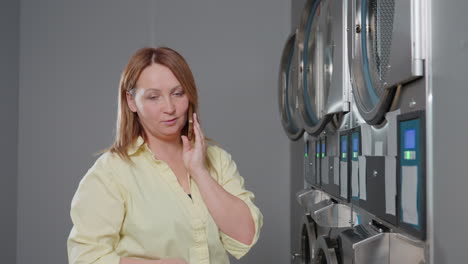 fair skinned lady stands near cloth dryer wall, speaks on phone during work inside laundromat, calm expression, stainless machines with blue screens in row, customer service coordination