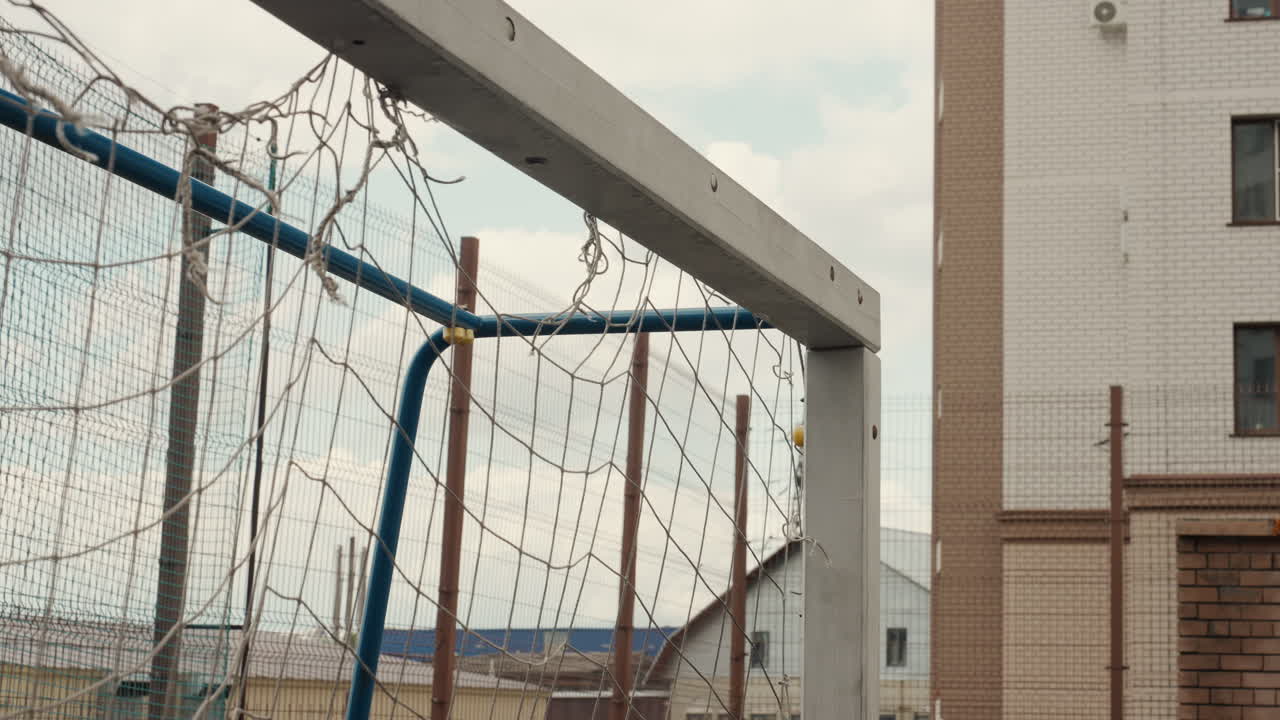 empty football goal drenched in rain, rusty metal frame and frayed net hang against residential building backdrop, cloudy sky and wet puddle on pitch creating melancholic, nostalgic urban atmosphere