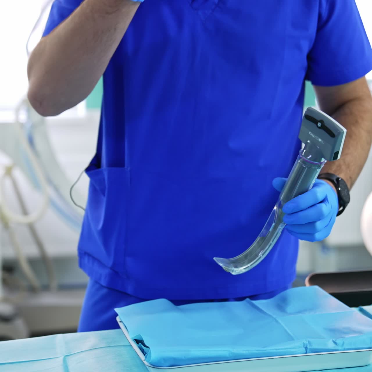 Anesthesiologist wearing bright blue uniform and latex gloves holding the portable video laryngoscope. Modern surgery room backdrop