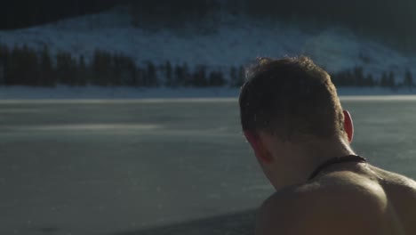 joven con pelo corto y bigote sentado sin camisa al lado de un lago de montaña congelado con nieve en el suelo