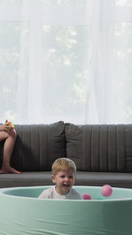 couple of kids spends time in living room. concentrated girl plays with rubik cube sitting on sofa and boy jumps into dry pool with balls laughing enthusiastically