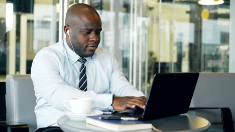 african american businessman in formal wear working and surfing social media on laptop while listening to music with earbuds in his ears in a glassy cafe