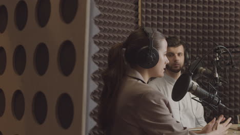 Eine-Elegante-Junge-Frau-Mit-Pferdeschwanz-Spricht-In-Einem-Radiostudio-über-Ein-Interessantes-Thema,-Während-Ein-Junger-Mann-Neben-Ihr-Zuhört