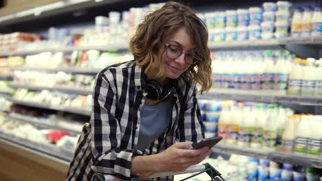 compras, tecnología, ventas, consumismo y concepto de personas - mujer con teléfono inteligente y auriculares en el cuello caminando en el supermercado. mujer sonriente enviando mensajes de texto con amigos, apoyada en su carrito de compras. vista lateral