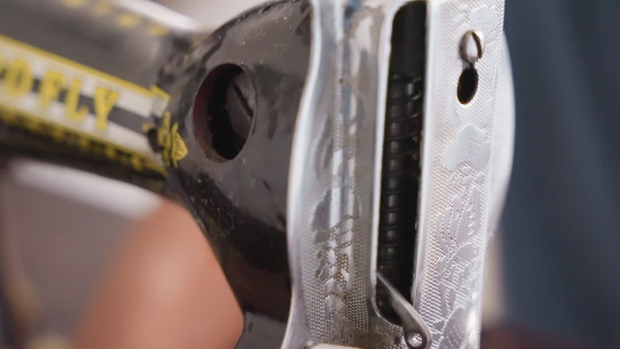 Nærbillede af sort maskinhoved i metal, emaljeret overflade med slidt logo, spoleåbning og spændingsåbning synlig, tekstureret maling og patina, nostalgisk værksteds- eller køkkenreparationsscene.