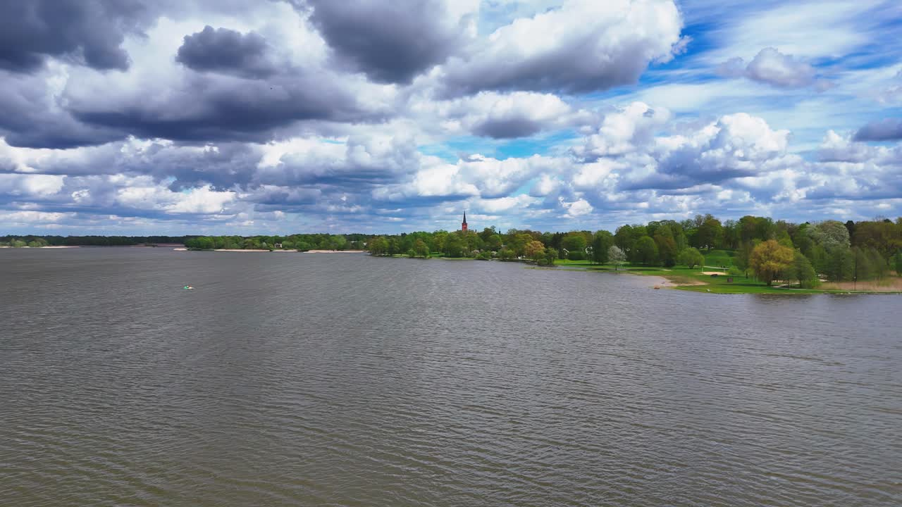 Aerial shot of Biržai's Lake Širvėna in late spring, featuring grey-blue waters under a dramatic sky with dark and white clouds, and a green shoreline with a distant red-roofed building and spire