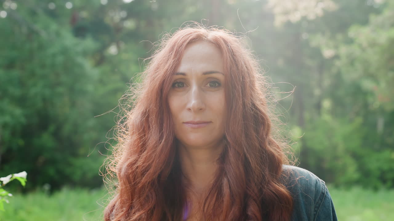Portrait of excited mother with long red hair smiling brightly in soft natural sunlight, standing in green forest background, joyful facial expression radiating warmth, and happiness outdoors