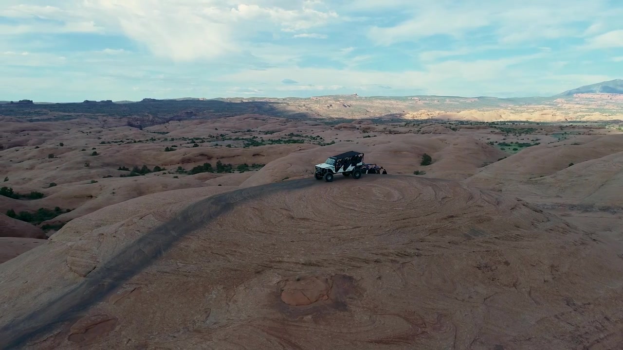 Una ripresa con drone 4K in tracking di una jeep rialzata che fa fuoristrada attraverso il paesaggio desertico estremo e roccioso vicino a Moab, Utah, con le Montagne Rocciose innevate che si stagliano in lontananza