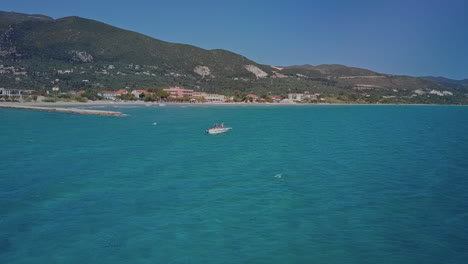 wide aerial as small boat bobs in blue water near shoreline of greek island