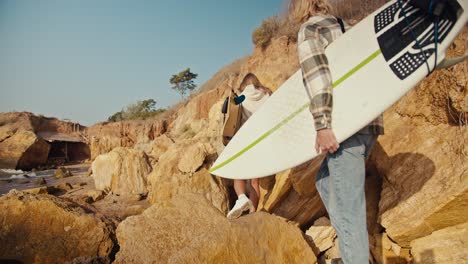 A-blond-guy-in-a-plaid-shirt-and-his-blond-girlfriend-in-a-white-sweatshirt-walk-along-a-rocky-beach-near-the-sea-and-carry-their-surfboards-with-them-on-a-sunny-morning