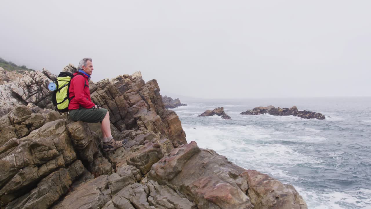 해변 근처에서 하이킹을 하는 동안 바위 위에 앉아 있는 배 ⁇ 을 가진 고령 하이커 남자.