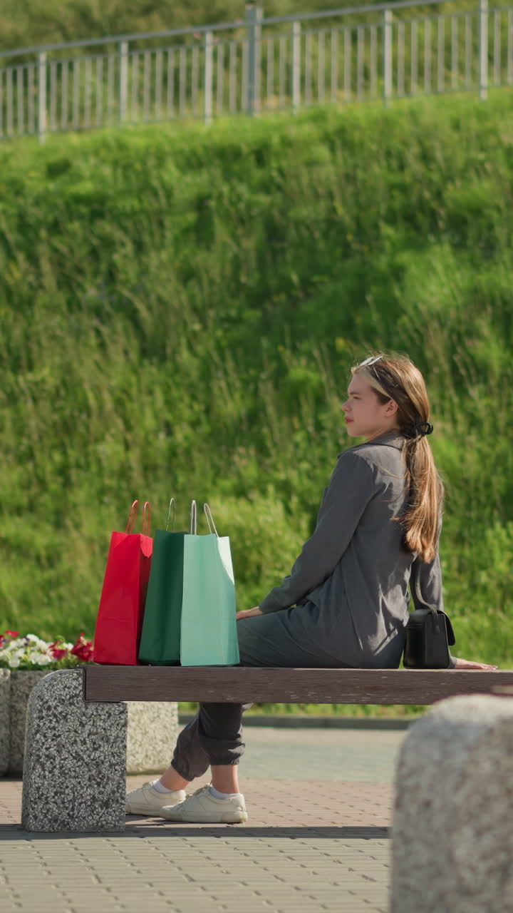 la dama camina hasta el banco, deja caer bolsas de compras de colores, se sienta, relaja la mano izquierda en el banco, ajusta la pierna y mira a la distancia, la vegetación y los postes de luz visibles en la colina en el fondo