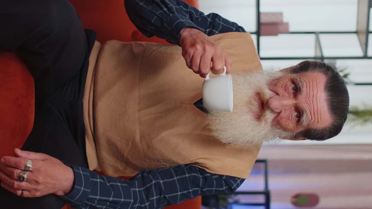 집 의 소파 에서 커피 나 허브 차 한 잔 을 마시는 매력적 인 미소 를 지닌 노인 할아버지