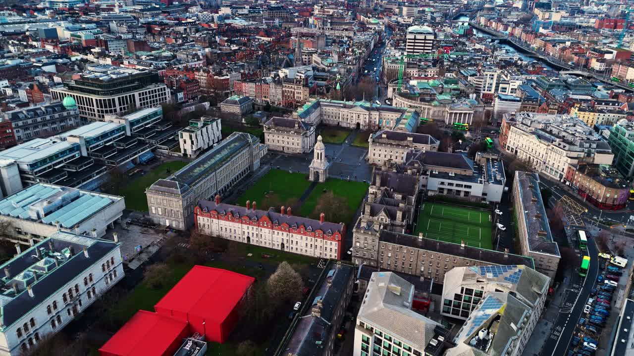 Lot z drona nad kampusem Trinity College w sercu Dublina o wschodzie słońca, gdzie studenci i turyści spacerują wśród pól i drzew.
