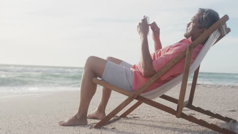 hombre latino senior relajándose en una cama de sol en la playa al atardecer, usando un teléfono inteligente