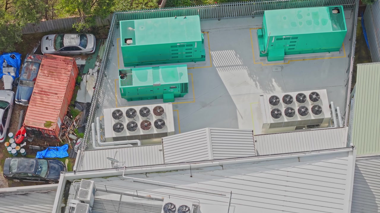 Drone shot of a secure data centre rooftop featuring industrial green generators, large HVAC units and fencing, under soft daylight. No people are visible, clean composition highlights facility.