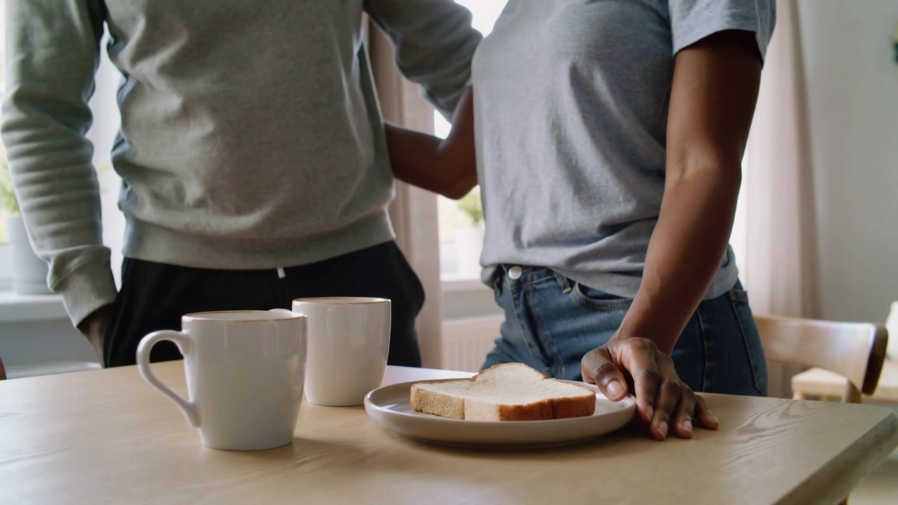 Moving man placing arm around woman's waist at kitchen table with mugs and bread, seeking quiet