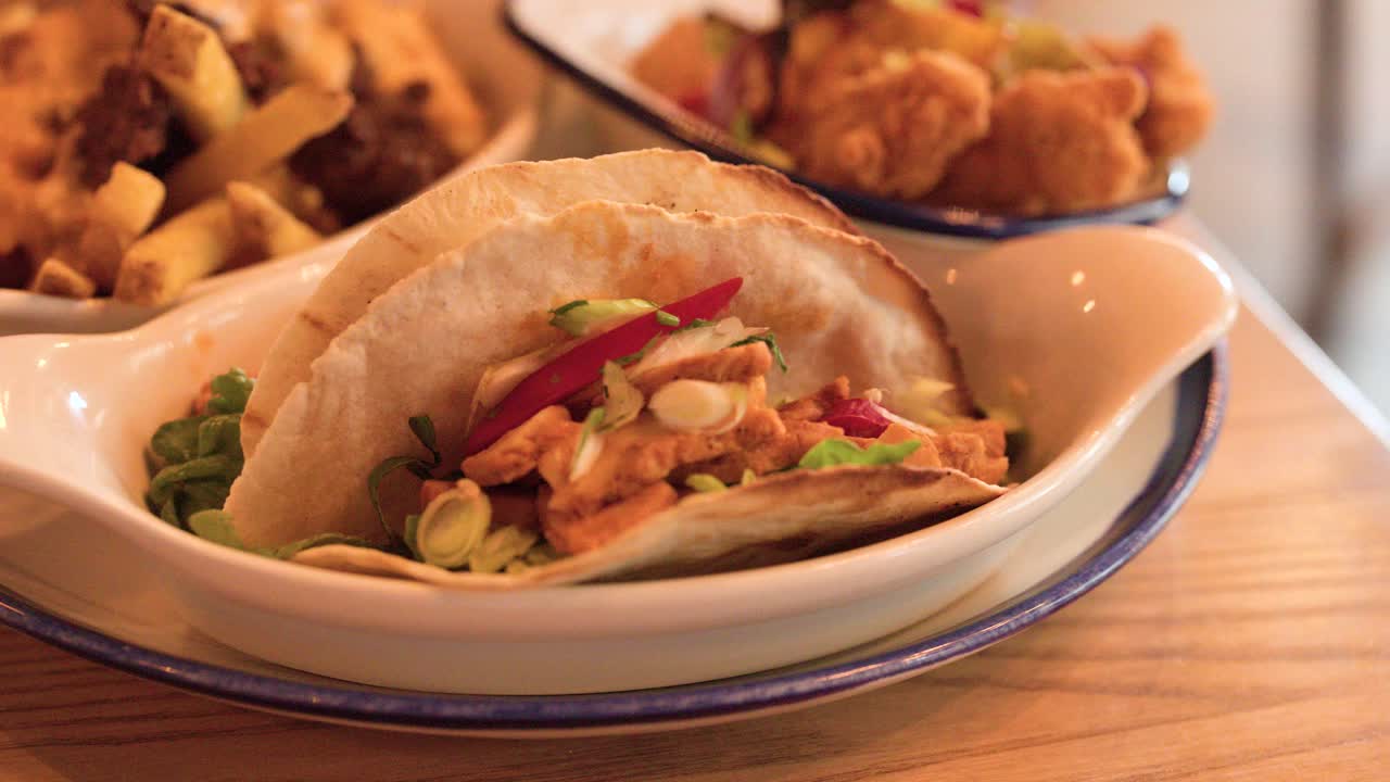 Hand serves plate of fried chicken tacos and cheesy loaded fries on wooden restaurant table