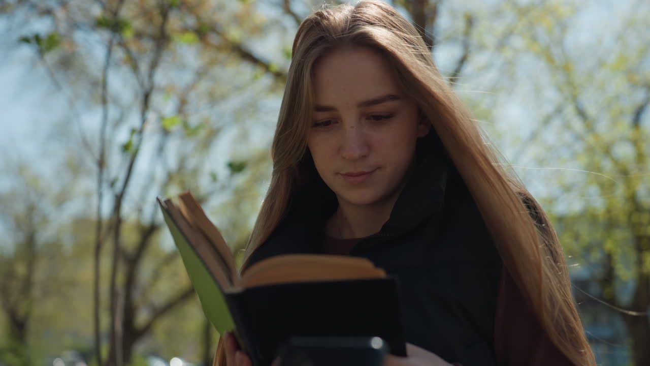Una mujer blanca lee sola bajo los árboles; sus gestos pensativos y su mano en el pelo transmiten una reflexión tranquila bajo la luz del sol primaveral; una escena íntima de estudio con un volumen clásico, pasando las páginas lentamente y con actitud contemplativa.