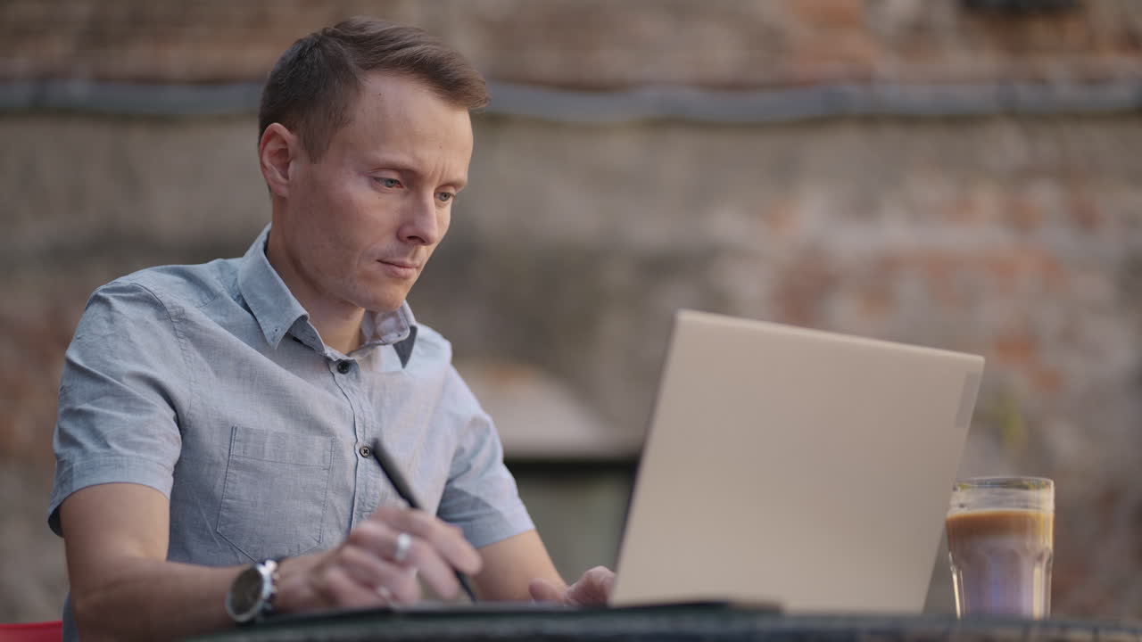 A man sitting in a street cafe works remotely writing a pen in a notebook. Work at the laptop. A man works remotely on a laptop