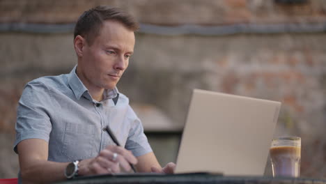 a man sitting in a street cafe works remotely writing a pen in a notebook. work at the laptop. a man works remotely on a laptop