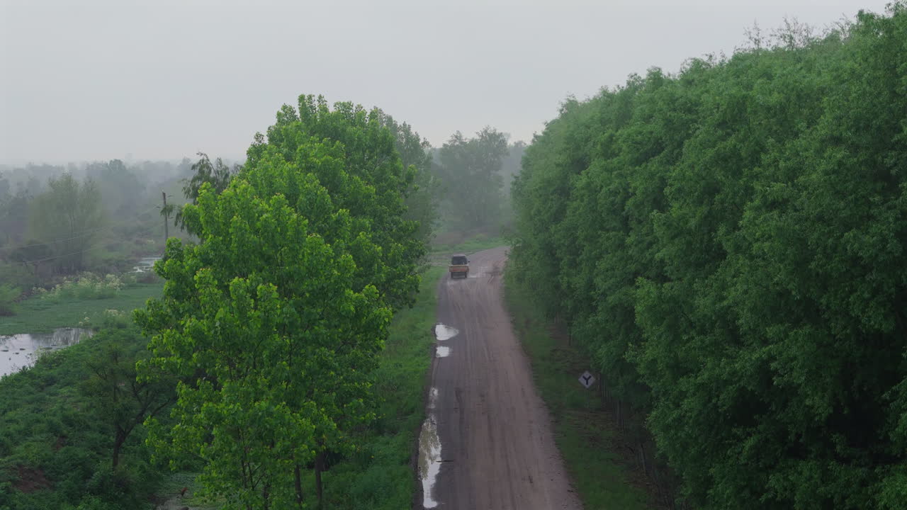 Aerial of an old car on a rural dirt road, surrounded by lush greenery and trees into misty wet forest path