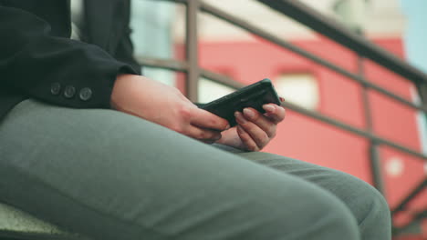 Side view of girl with artificial nails using smartphone while seated outdoors in urban space, wearing grey pants, with iron railing and modern architecture in softly blurred background