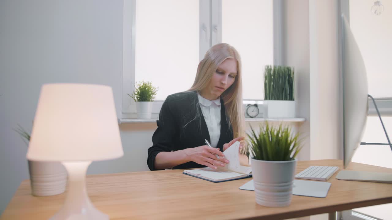 Bella giovane donna bionda seduta alla scrivania che scrive con una penna su un blocco note, girando una pagina e strappando fogli dal suo quaderno.