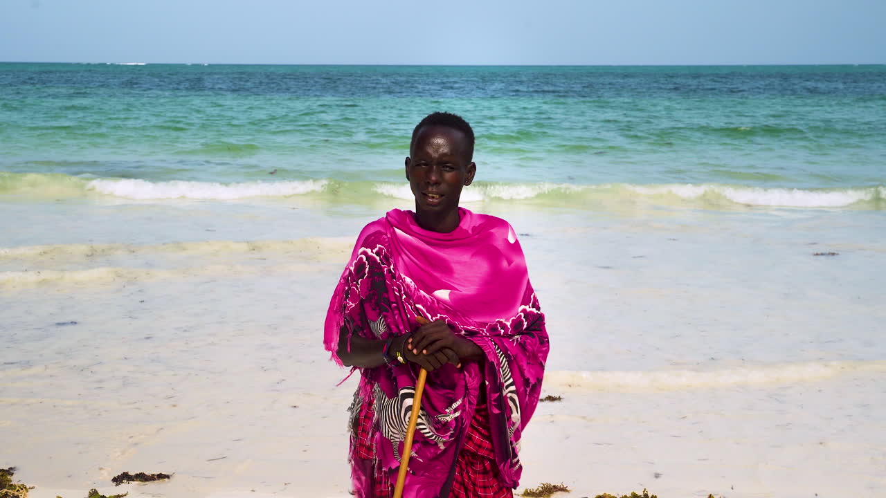 Uomo Maasai africano con abiti rosa in piedi sulla spiaggia, appoggiato a un bastone.