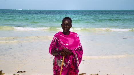 african maasai man in pink clothing standing on beach leaning on stick