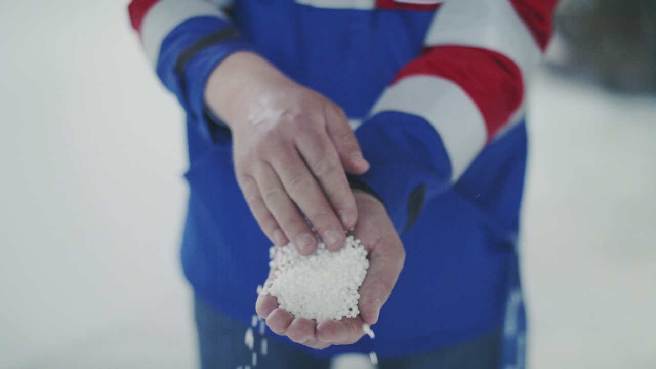A medium close-up shot shows a person's hands pouring white, round pellets from one hand to another in slow motion. The product could be fertilizer, seeds, or de-icing salt