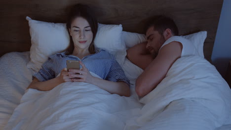 joven mujer caucásica con cabello oscuro acostado en la cama, charlando en el teléfono inteligente a altas horas de la noche y mirando a su esposo que duerme a su lado
