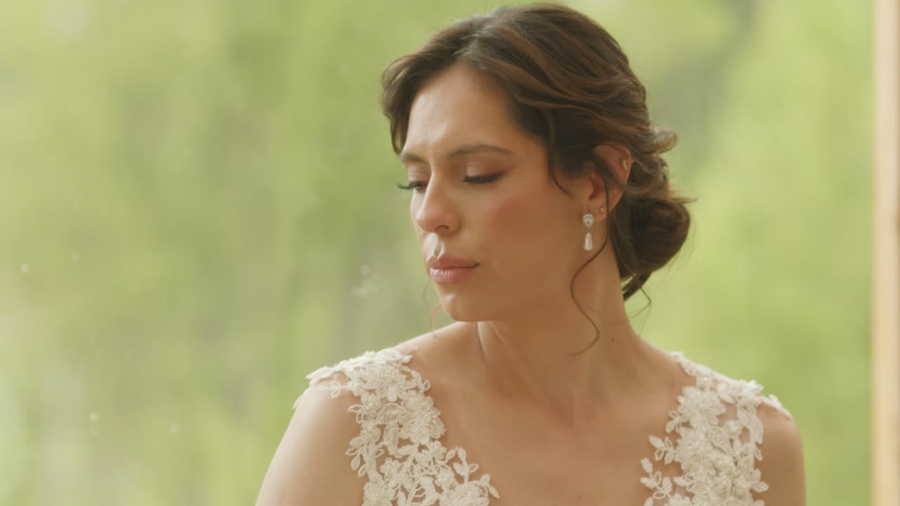Medium shot of a bride looking to the side, her expression calm and dreamy. The background is softly blurred, highlighting the beauty of the moment and the detail of her wedding dress.