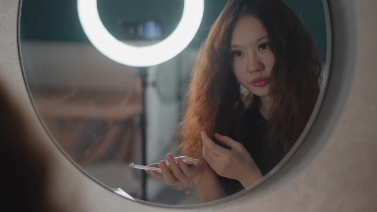 Giovane donna siede davanti a uno specchio rotondo illuminato da una luminosa luce ad anello in un camerino, ispezionando le unghie, sistemandosi i capelli e controllando il telefono durante una tranquilla routine di bellezza con un'atmosfera di cura di sé concentrata.