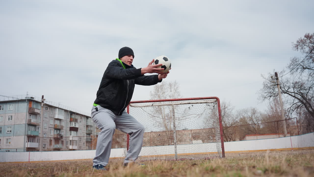 골키퍼가 웅크리고 앉아 축구공을 잡고 있어요. 백인 남성 키퍼가 흐린 날씨 속 도시 축구장에서 녹슨 골대 앞에서 낮은 자세로 막는 연습을 하고 있어요. 집중된 자세와 안정적인 손은 회복력과 준비성을 보여줍니다.