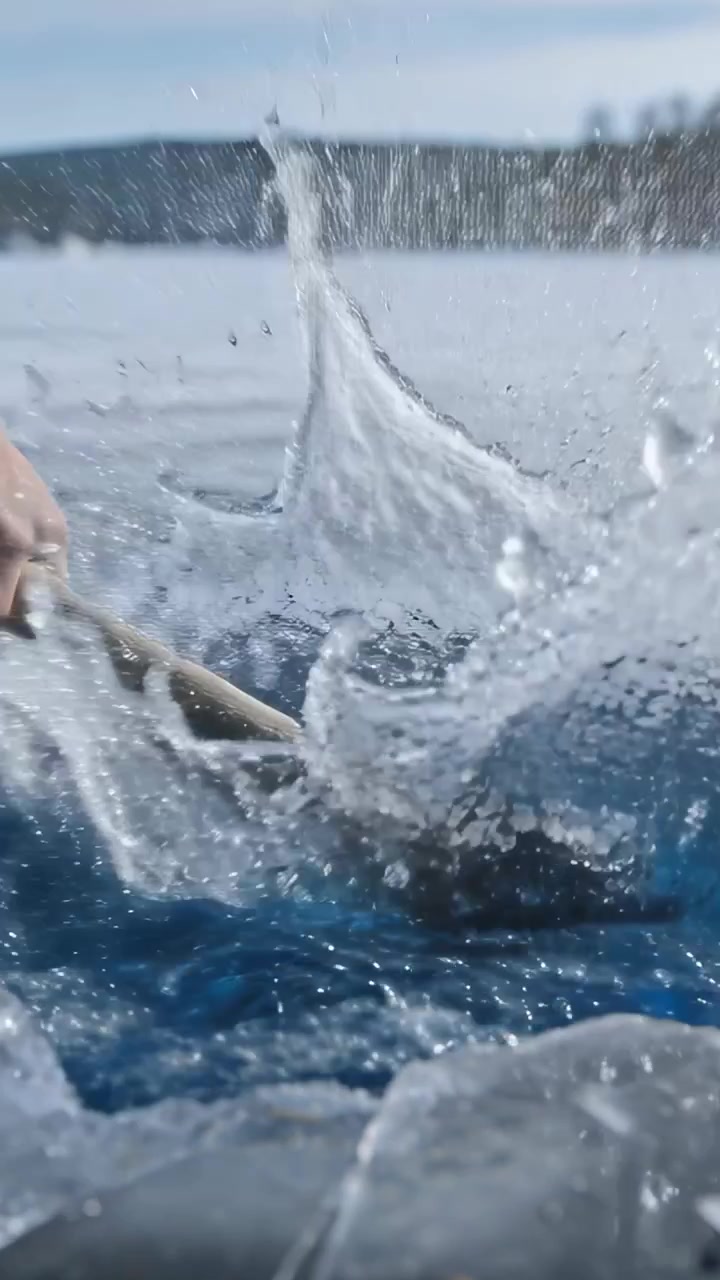 Video verticale: Ghiaccio che si rompe, mano nuda che impugna un'ascia di legno, tagliando un buco, esponendo acqua turchese.