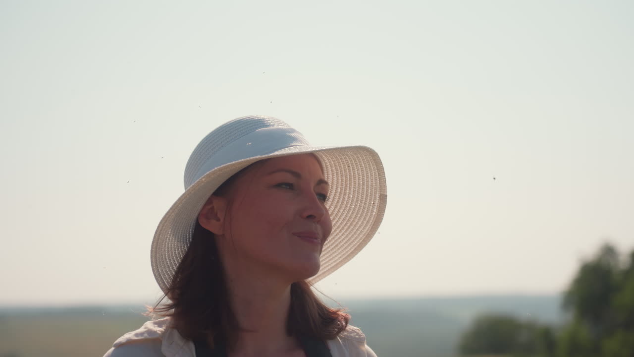 Close up of woman in white hat and cream shirt walking in countryside under bright sun, eyes closed with relaxed face, soft breeze gently moving hair, blurry rural landscape in distant background