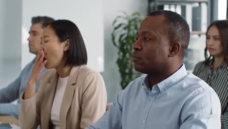 attendees in a contemporary office listen intently during a business presentation. body language indicates varying levels of engagement, with one person appearing tired.