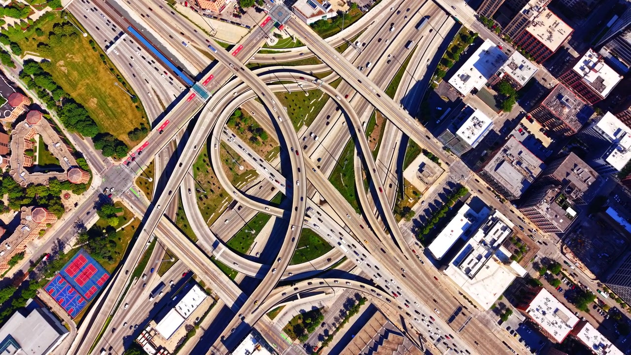 Complex highway interchange in city, Chicago. Overhead drone view of intersecting concrete roads, moving cars, green patches and nearby commercial buildings.