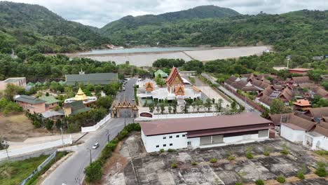 Drone footage captures a serene Buddhist temple surrounded by lush greenery and mountains in Phuket, Thailand, under soft daylight